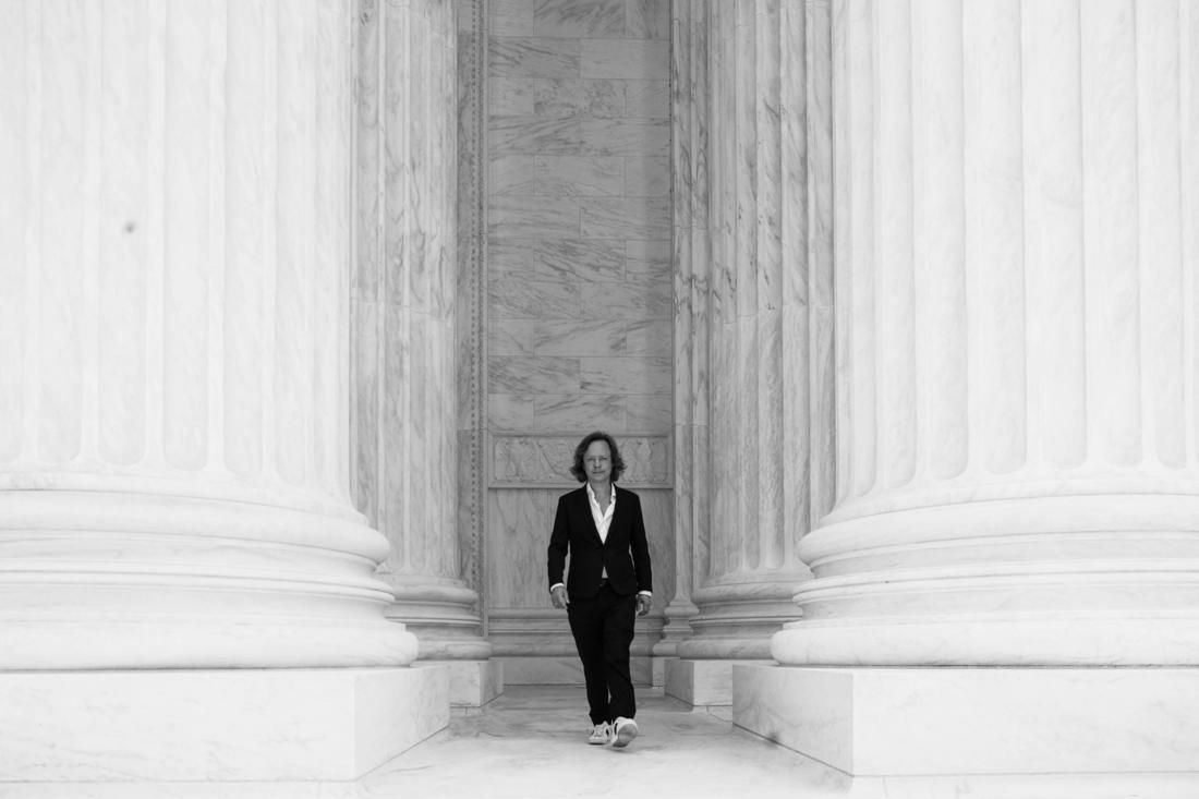 Brock Pierce walking out between tall marble columns, in black and white